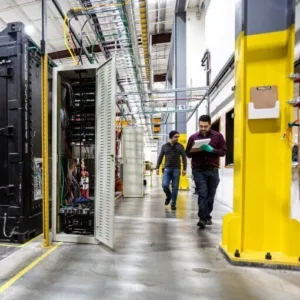 Two men walk toward camera in a long corridor near lots of equipment and industrial stuff