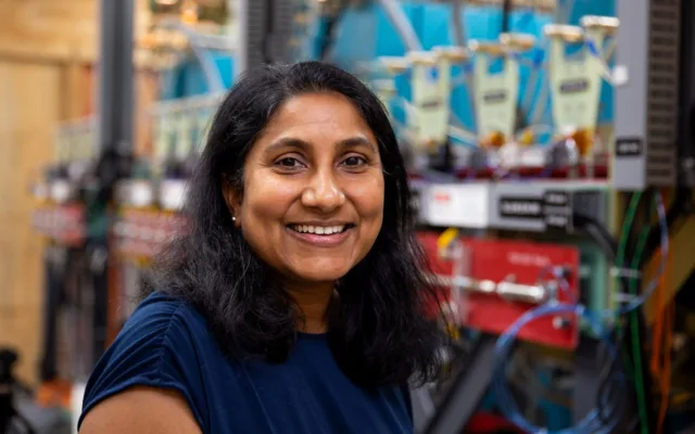 Photo of woman with dark hair and skin in a navy blue blouse smiling at the camera while standing in front of a large piece of electrical hardware
