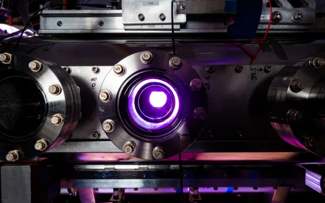 Purple light glows through a window looking into a stainless steel chamber