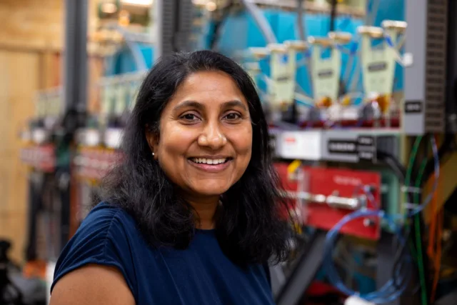 Photo of woman with dark hair and skin in a navy blue blouse smiling at the camera while standing in front of a large piece of electrical hardware
